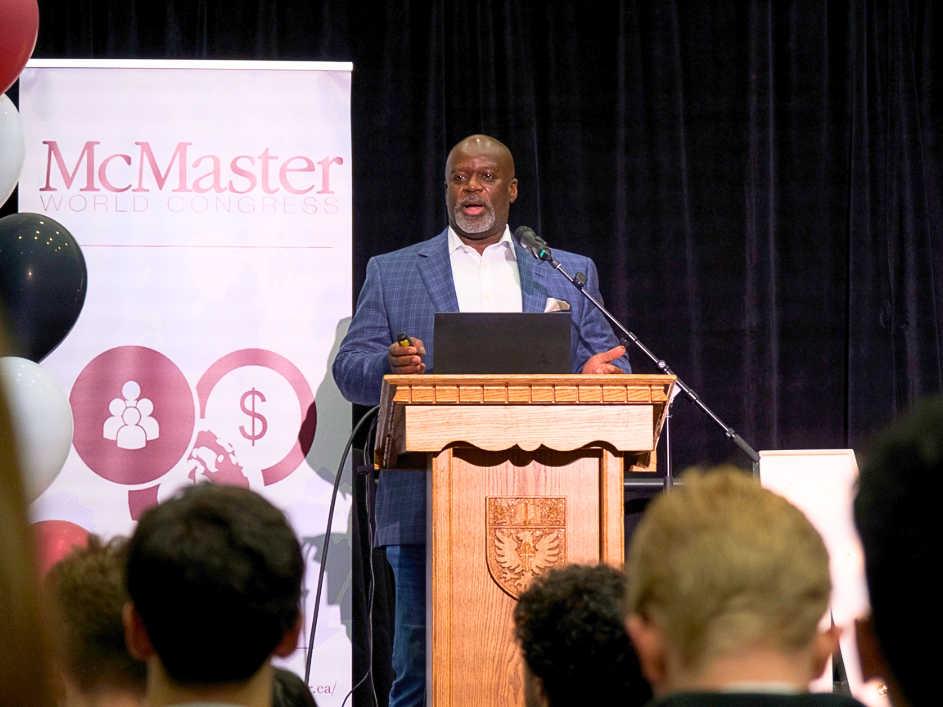A speaker stands at a podium, presenting to an attentive audience in a large venue.