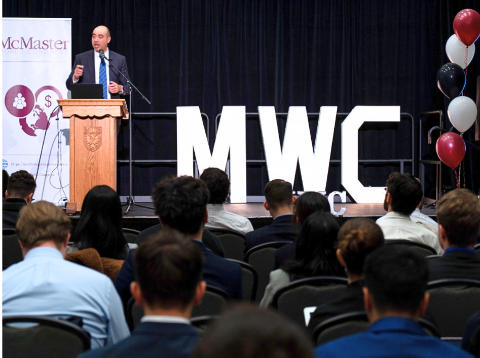 A man in a suit speaks at a podium beside a large sign that reads 