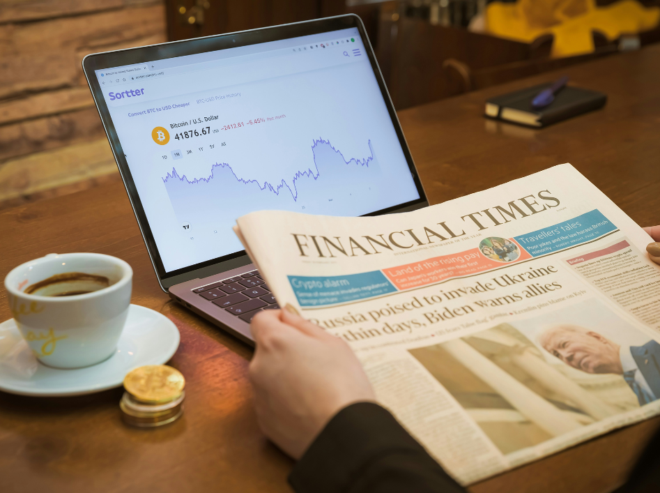 A person holding a newspaper on a table with a laptop displaying a stock graph and a coffee cup (left). The newspaper reads Financial Times.