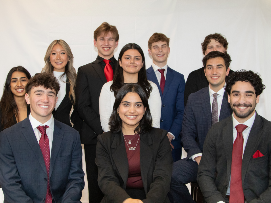 A group of ten young professionals dressed in formal business attire posing together in front of a white backdrop. They are smiling and arranged in two rows, with some seated in front and others standing behind.